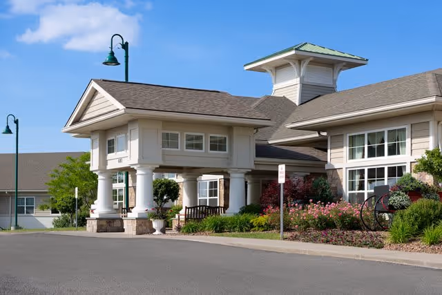 Exterior view of a senior living facility building with a covered entrance supported by white columns, surrounded by well-maintained landscaping including bushes and flowers, under a clear blue sky.
