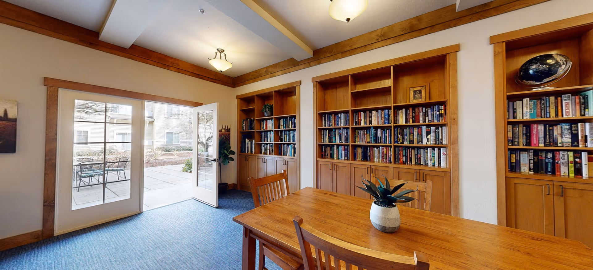 A cozy room with wooden bookshelves filled with books along the wall, a wooden table with chairs around it, and a potted plant on the table. French doors open to an outdoor patio area with a table and chairs. The room has a blue carpet and warm lighting from ceiling fixtures.