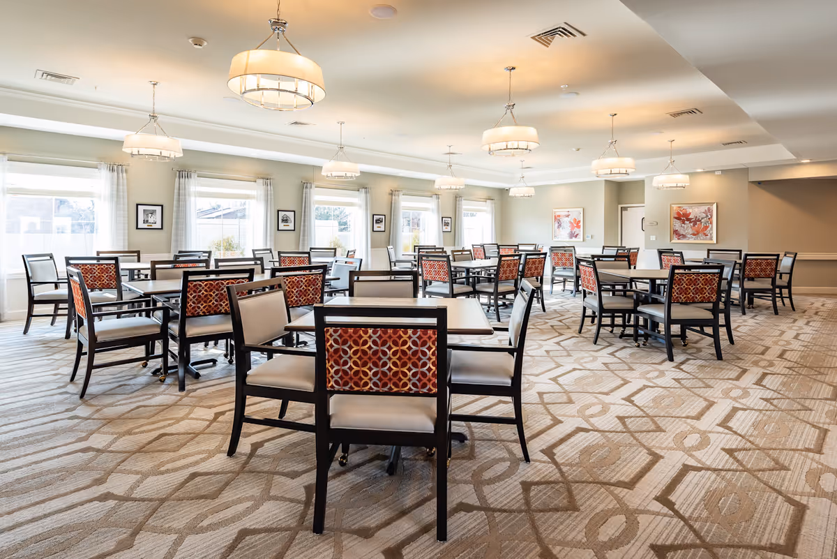 A spacious dining room with multiple square tables and chairs arranged neatly. The chairs have a patterned red and orange backrest with beige seats. The room is well-lit with large windows covered by sheer white curtains and several modern pendant lights hanging from the ceiling. The carpet has a geometric pattern in neutral tones, and framed artwork is displayed on the walls.