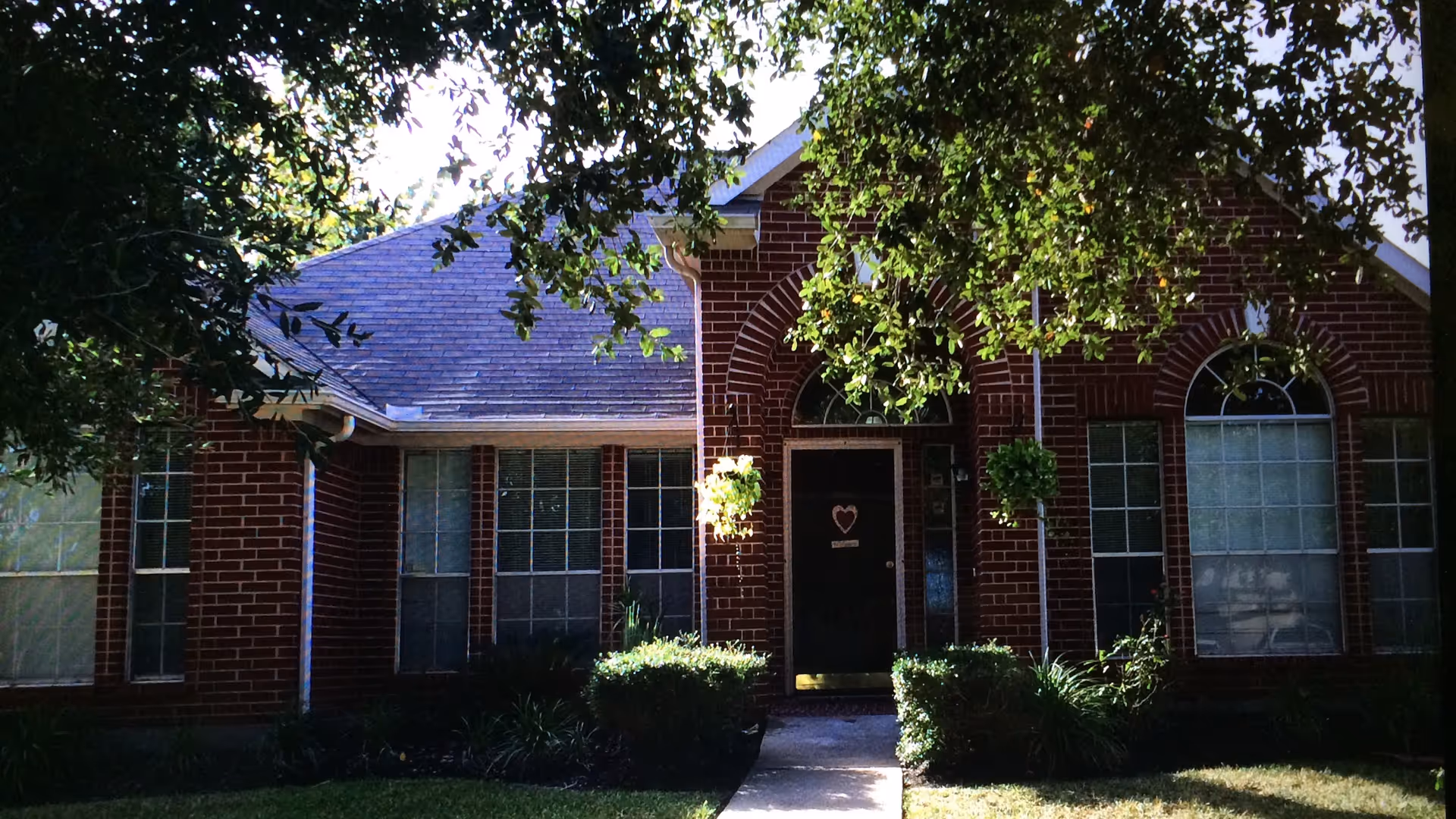 Front exterior of a red-brick single-story house with arched windows, a central dark front door decorated with a heart, hanging plants, and shrubs under large overhanging trees.