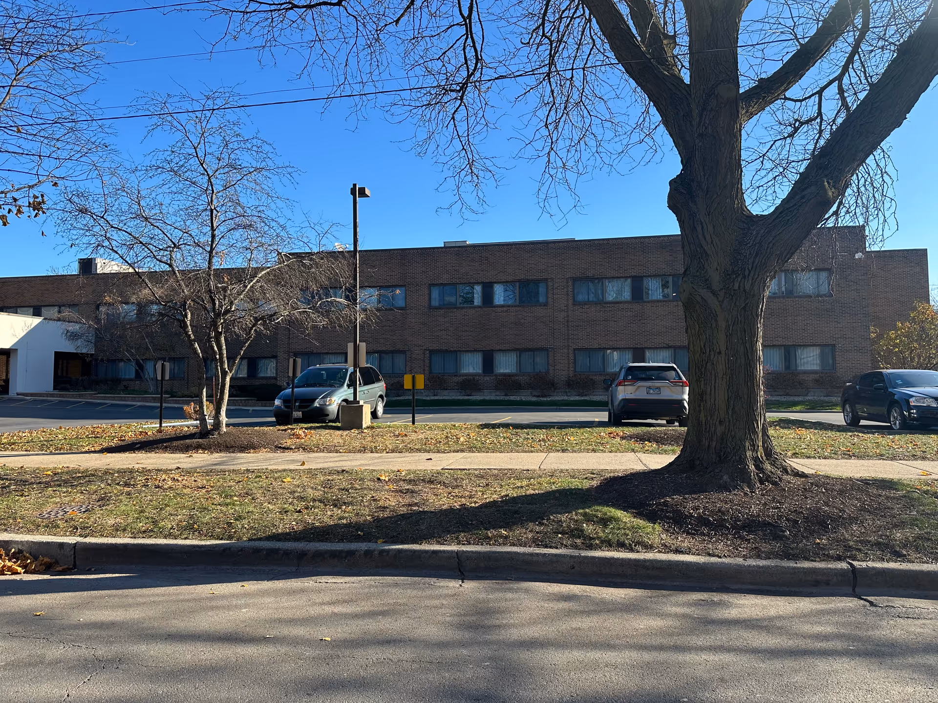 Brick two-story senior living facility building with parked cars and leafless trees in front under a clear blue sky.