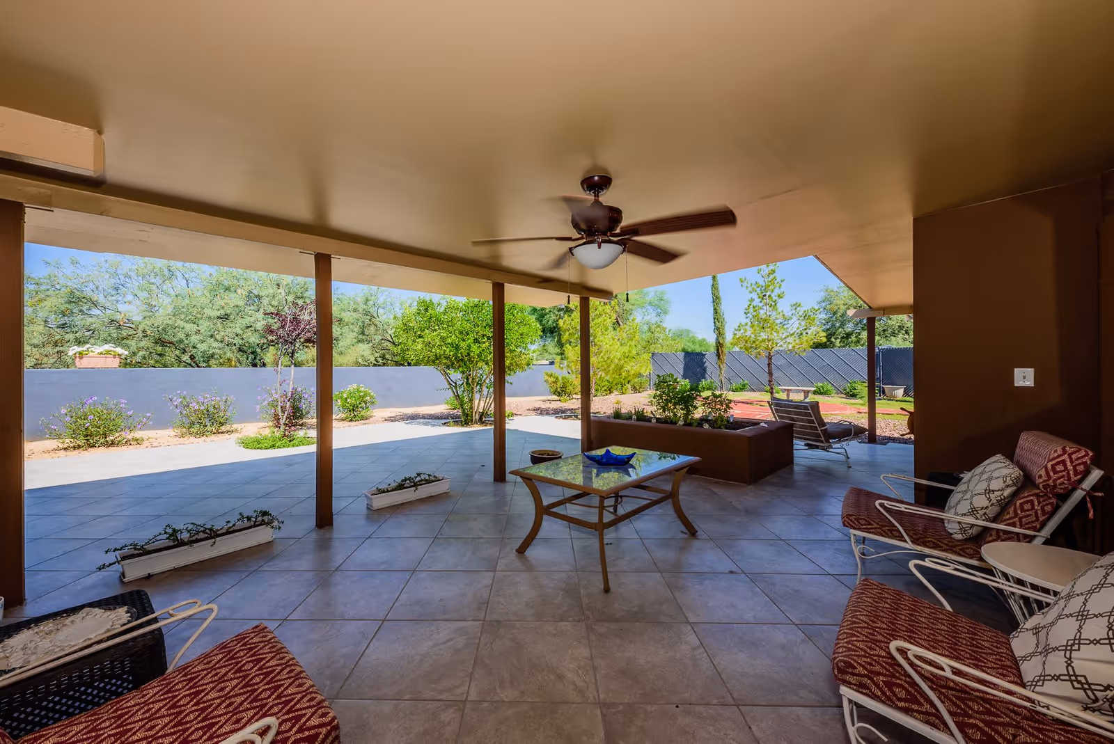 Covered outdoor patio with a ceiling fan, tiled floor, seating and a table overlooking a landscaped yard.