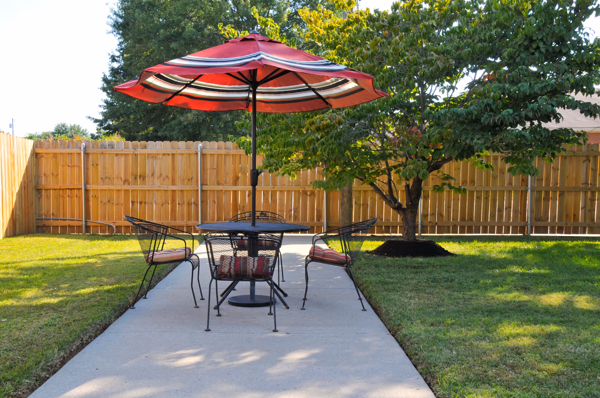 Patio with a round table, four metal chairs and a red striped umbrella on a concrete path in a fenced backyard with a tree.
