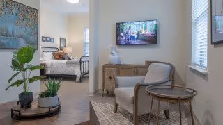 A cozy interior view of a senior living facility showing a small sitting area with a cushioned chair and round wooden side table, a wooden cabinet with a decorative vase, and a wall-mounted TV displaying a nature scene. In the background, a bedroom with a neatly made bed and framed artwork on the wall is visible. The room is decorated with plants and has large windows letting in natural light.