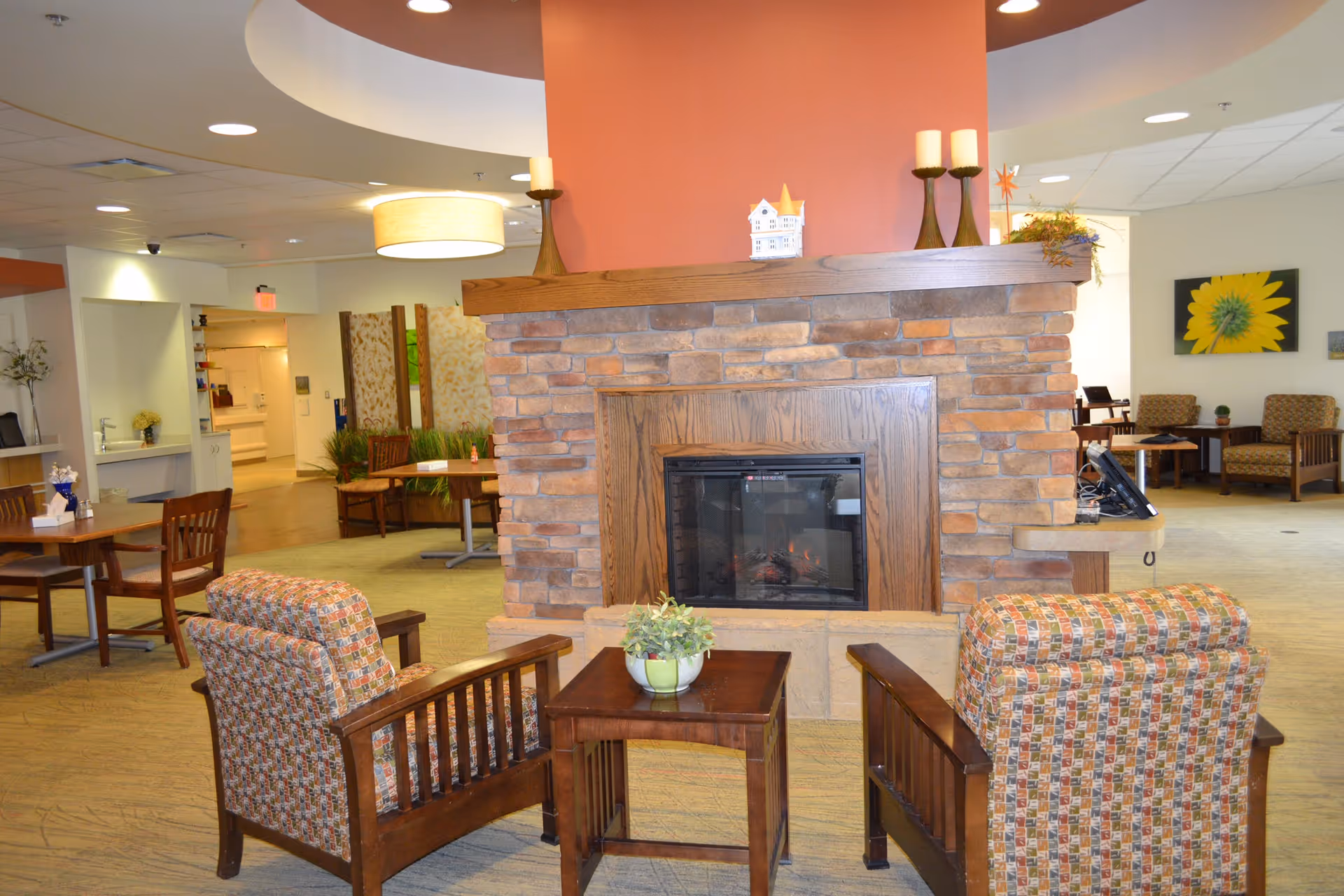 A cozy common area in a senior living facility featuring a stone fireplace with a wooden mantle, two patterned armchairs facing the fireplace, and a small wooden table with a potted plant between them. In the background, there are additional seating areas with chairs and tables, warm lighting, and decorative elements including candles and a sunflower painting on the wall.