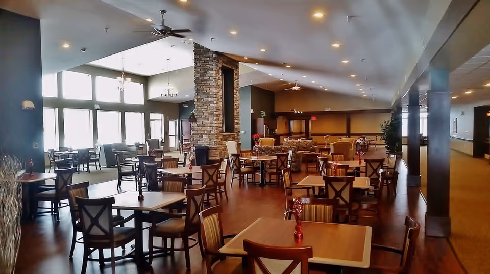 A spacious dining room with multiple wooden tables and chairs arranged neatly. The room features a stone fireplace in the center, large windows letting in natural light, ceiling fans, and warm lighting from recessed ceiling lights. There are also some comfortable armchairs and sofas in the background near the fireplace.
