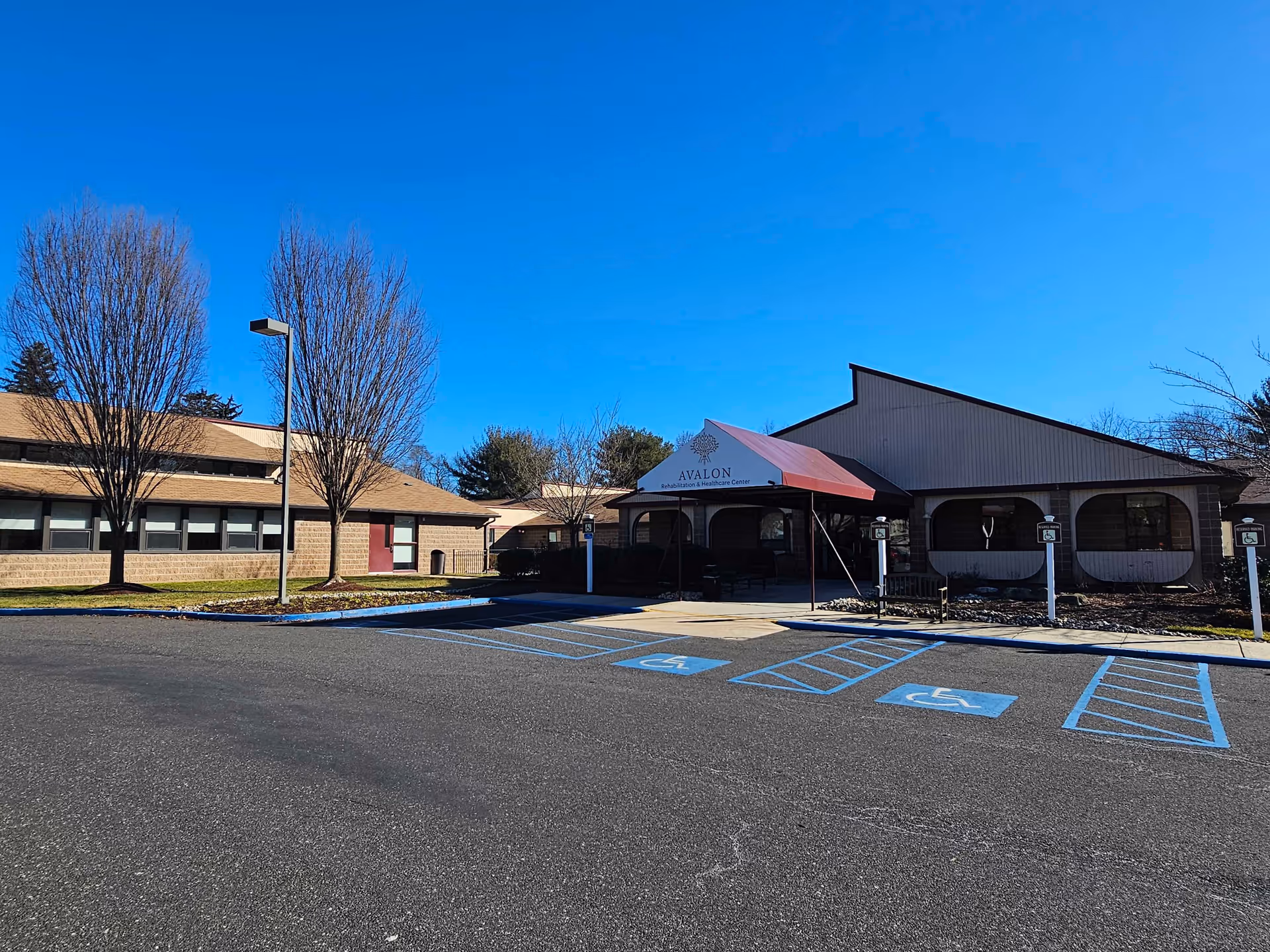 Front exterior of Avalon Rehabilitation & Healthcare Center with a covered entrance, handicap parking spaces, and leafless trees under a clear blue sky.