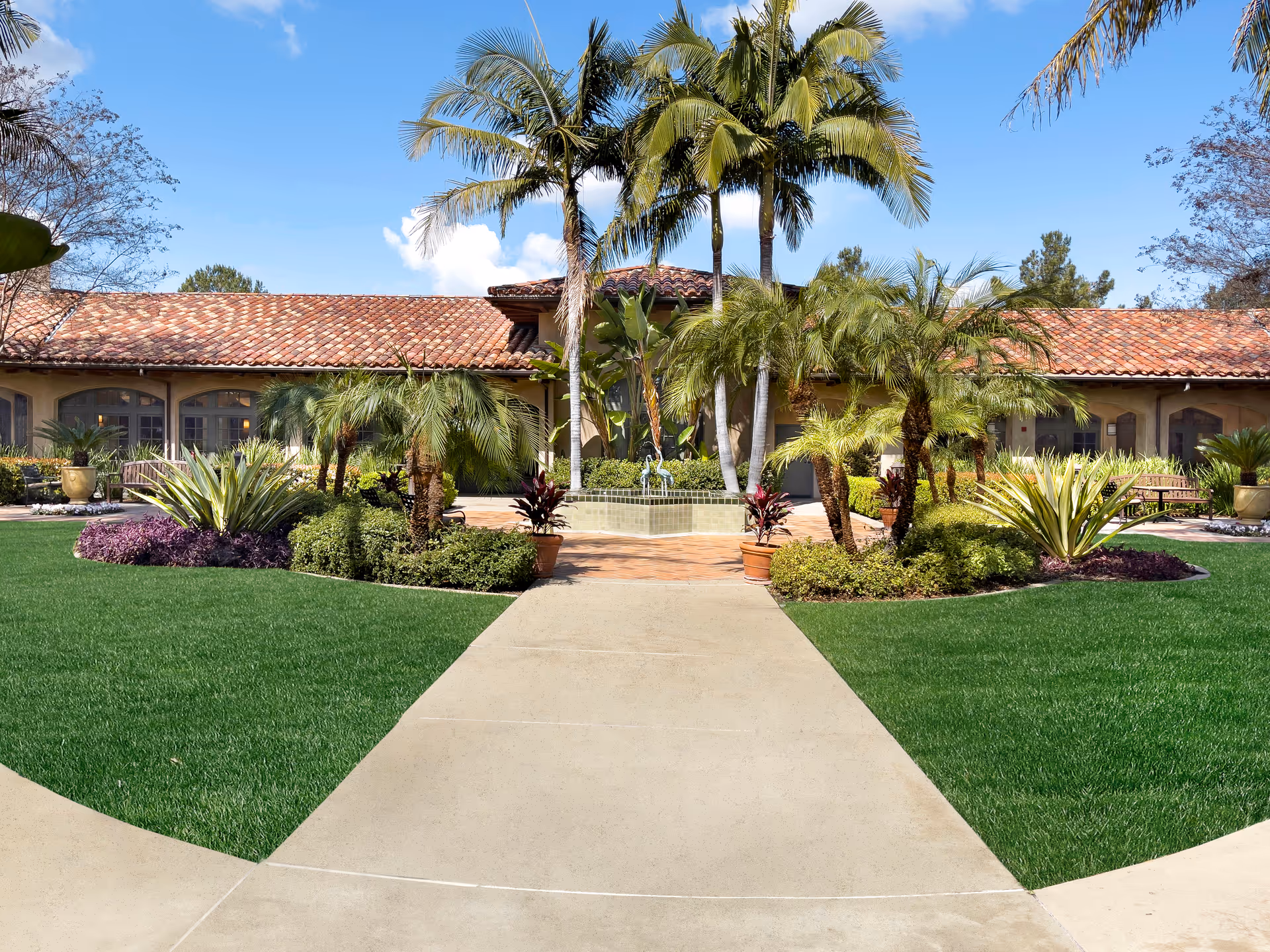 Outdoor view of a senior living facility with a tiled roof building in the background, surrounded by palm trees, various plants, and well-maintained green lawns. A concrete pathway leads to the entrance area with a small fountain and benches visible.