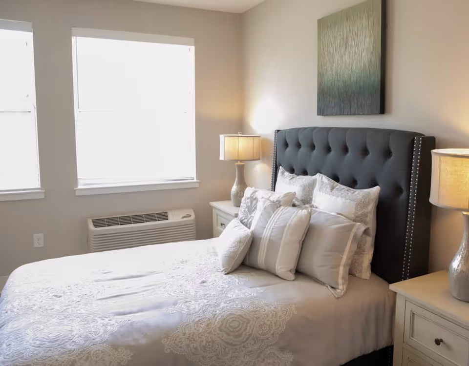 Well-lit bedroom with a tufted dark headboard, decorative pillows, bedside tables and lamps beneath two windows.