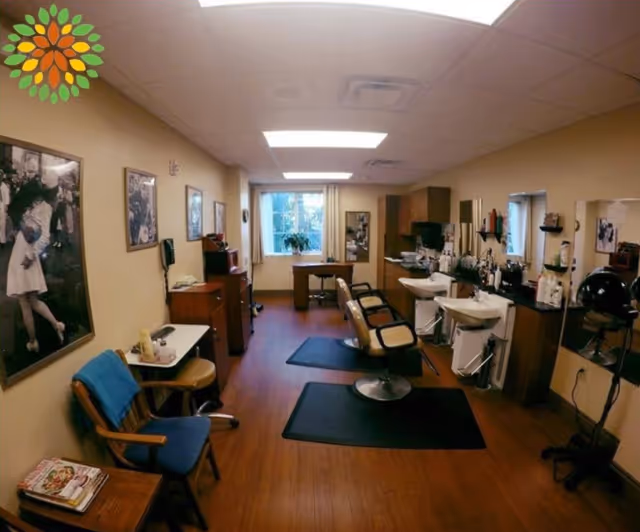 Interior view of a salon area in a senior living facility with two salon chairs in front of sinks, a hair dryer, wooden flooring, and framed black and white photos on the walls. There are also chairs and small tables along the left wall and a window at the far end letting in natural light.
