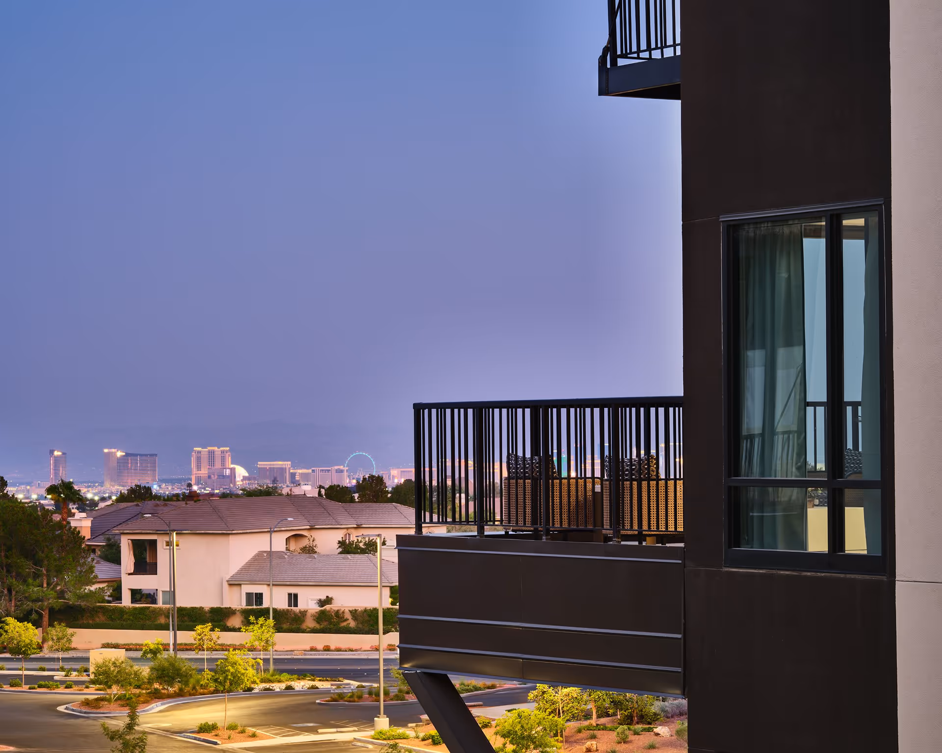 View of a balcony with outdoor seating on a modern building overlooking a suburban area with houses, trees, and a distant city skyline at dusk.