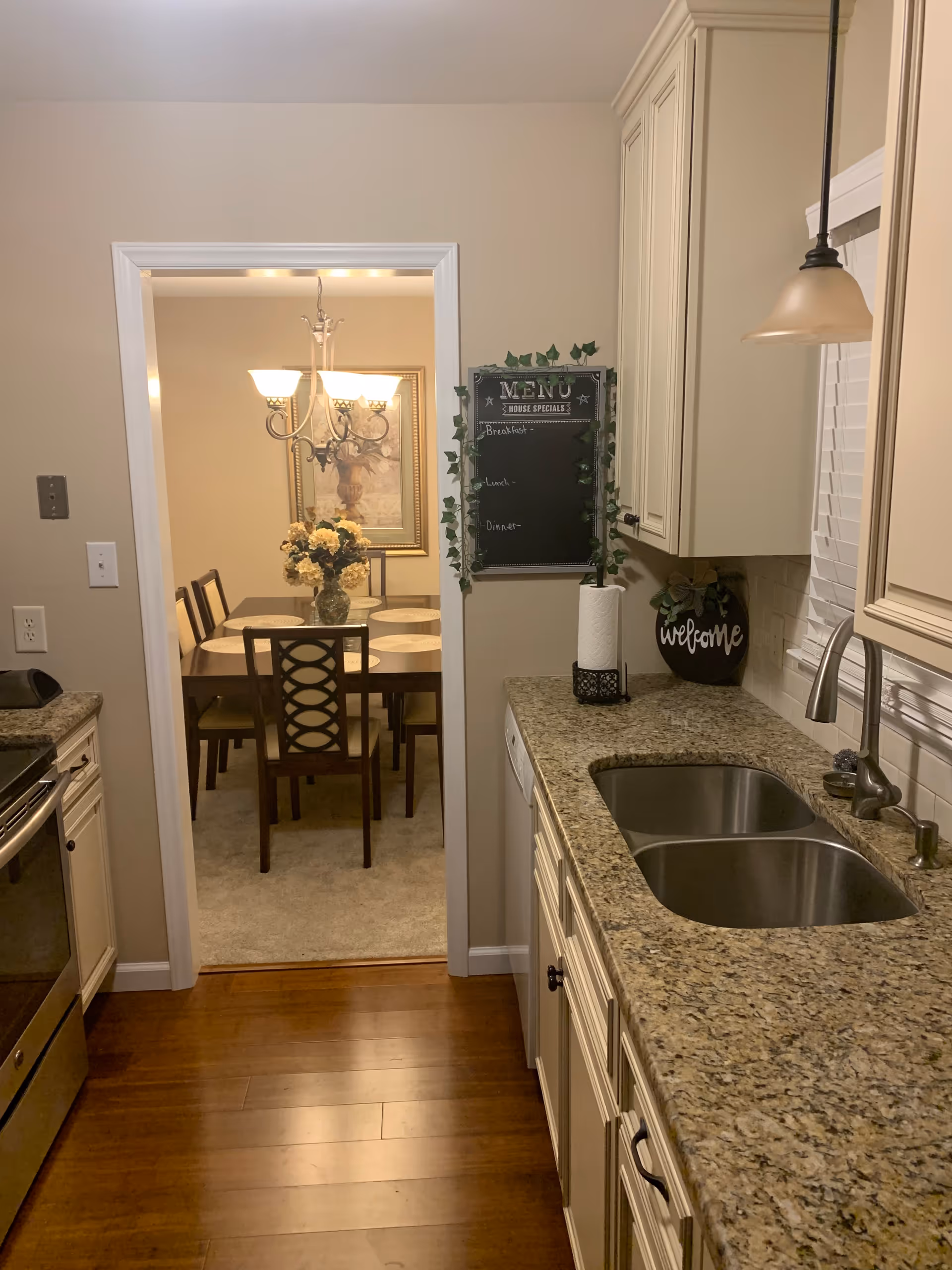 View down a kitchen with granite countertops and a double sink looking into a dining room with a chandelier and table.