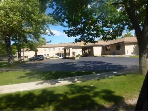 Exterior view of a single-story senior living facility building with a covered entrance and a parking area in front. The building is surrounded by green grass and large trees providing shade under a partly cloudy sky.