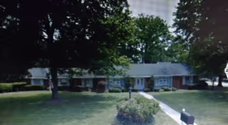 Single-story residential building with a gray roof and brick exterior, surrounded by green trees and a well-maintained lawn with a walkway leading to the front door.