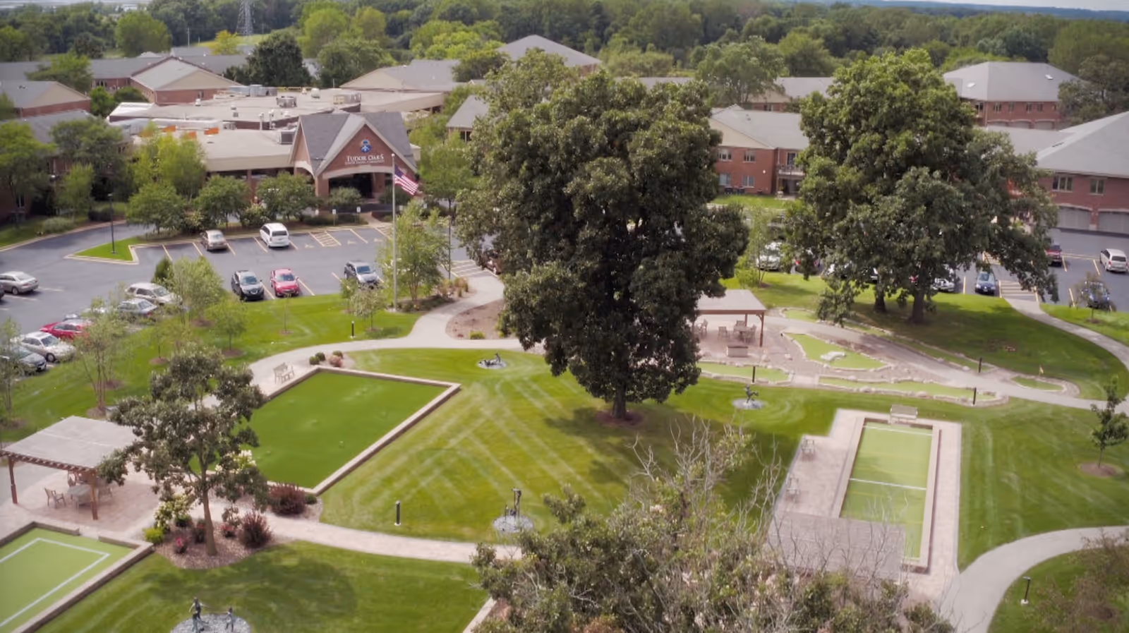 Aerial view of Tudor Oaks Senior Living Community showing the main building, parking lot, lawns, trees, and walking paths.