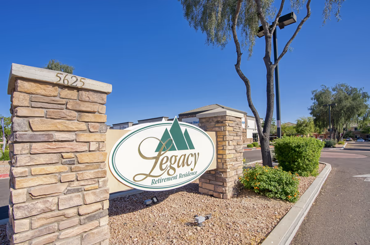 Stone pillars with the address number 5625 and a large oval sign reading 'Legacy Retirement Residence' at the entrance of a retirement community, with trees, bushes, and a clear blue sky in the background.