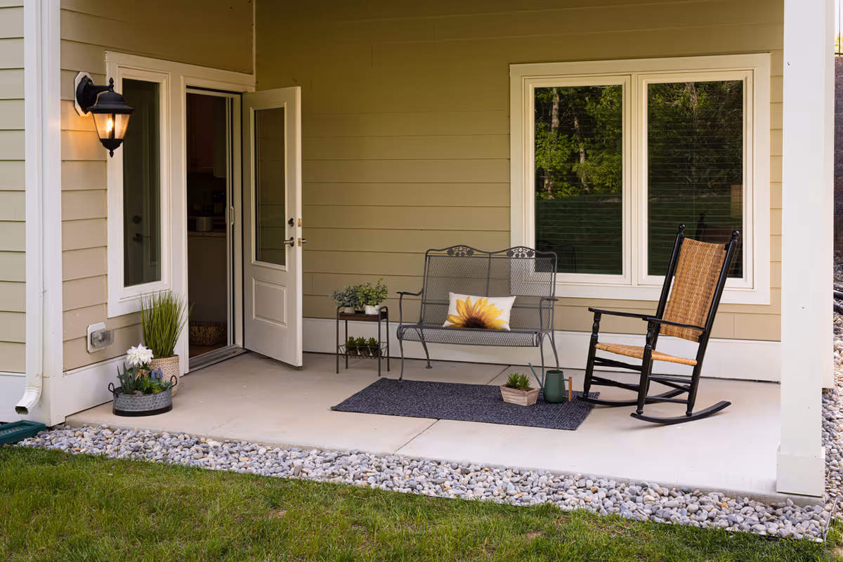 Covered patio with an open door, a metal bench with a sunflower pillow, a wicker rocking chair, potted plants, and a doormat.