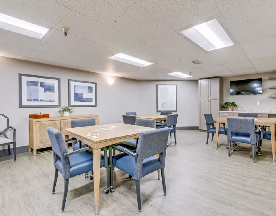 Bright communal dining/activity room with several wooden tables and blue upholstered chairs, wall art, cabinets and a mounted TV.