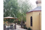 Outdoor patio area with tables and chairs under umbrellas next to a yellow building with arched windows, surrounded by trees and greenery.