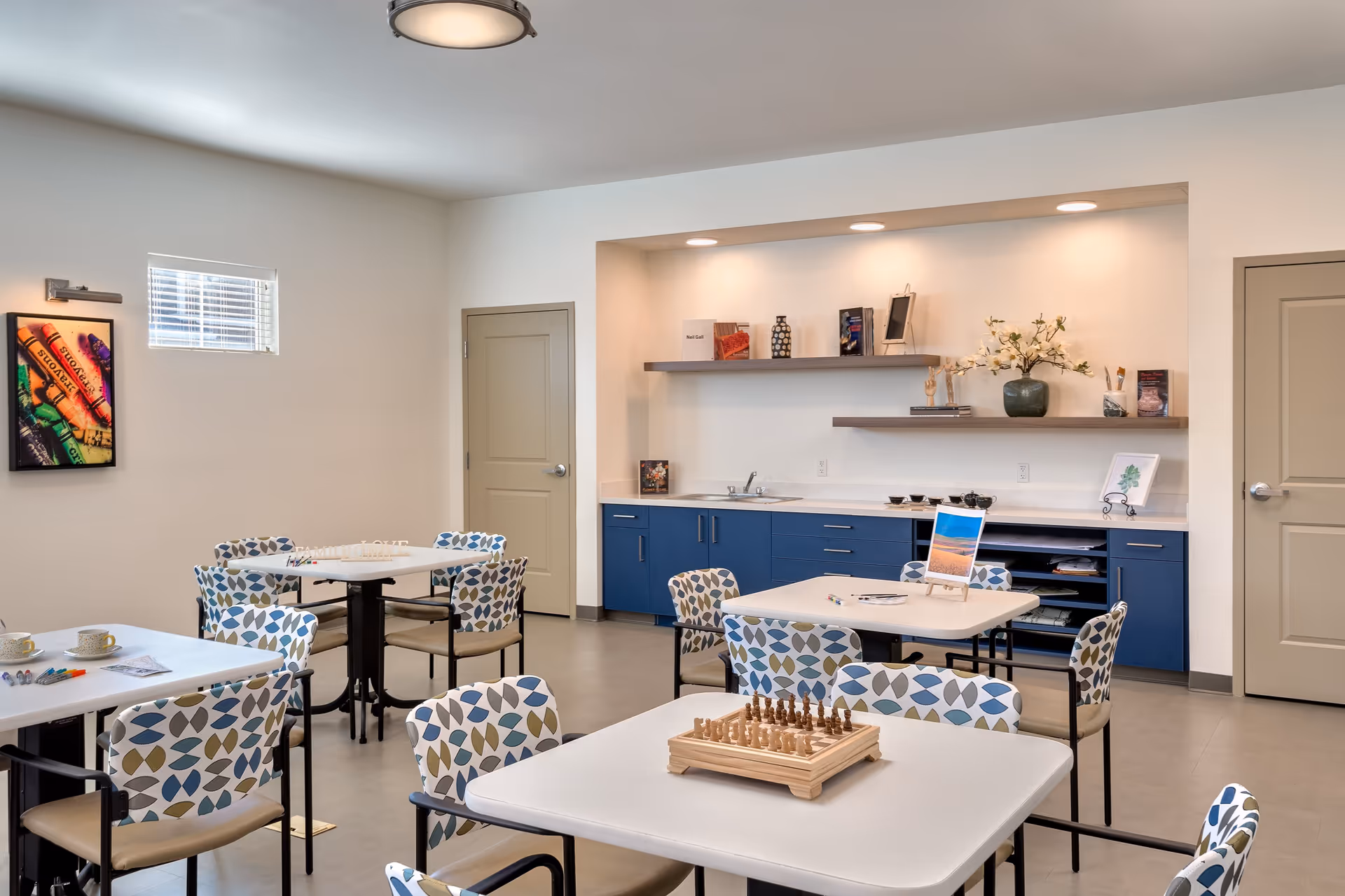 A bright and clean activity room with several tables and chairs featuring patterned upholstery. One table has a chessboard set up, another has art supplies and a small easel with a painting. The back wall has blue cabinets with a countertop, a small sink, and two floating shelves decorated with books, vases, and flowers. The room has neutral walls, two closed doors, a small window, and a colorful framed picture on one wall.