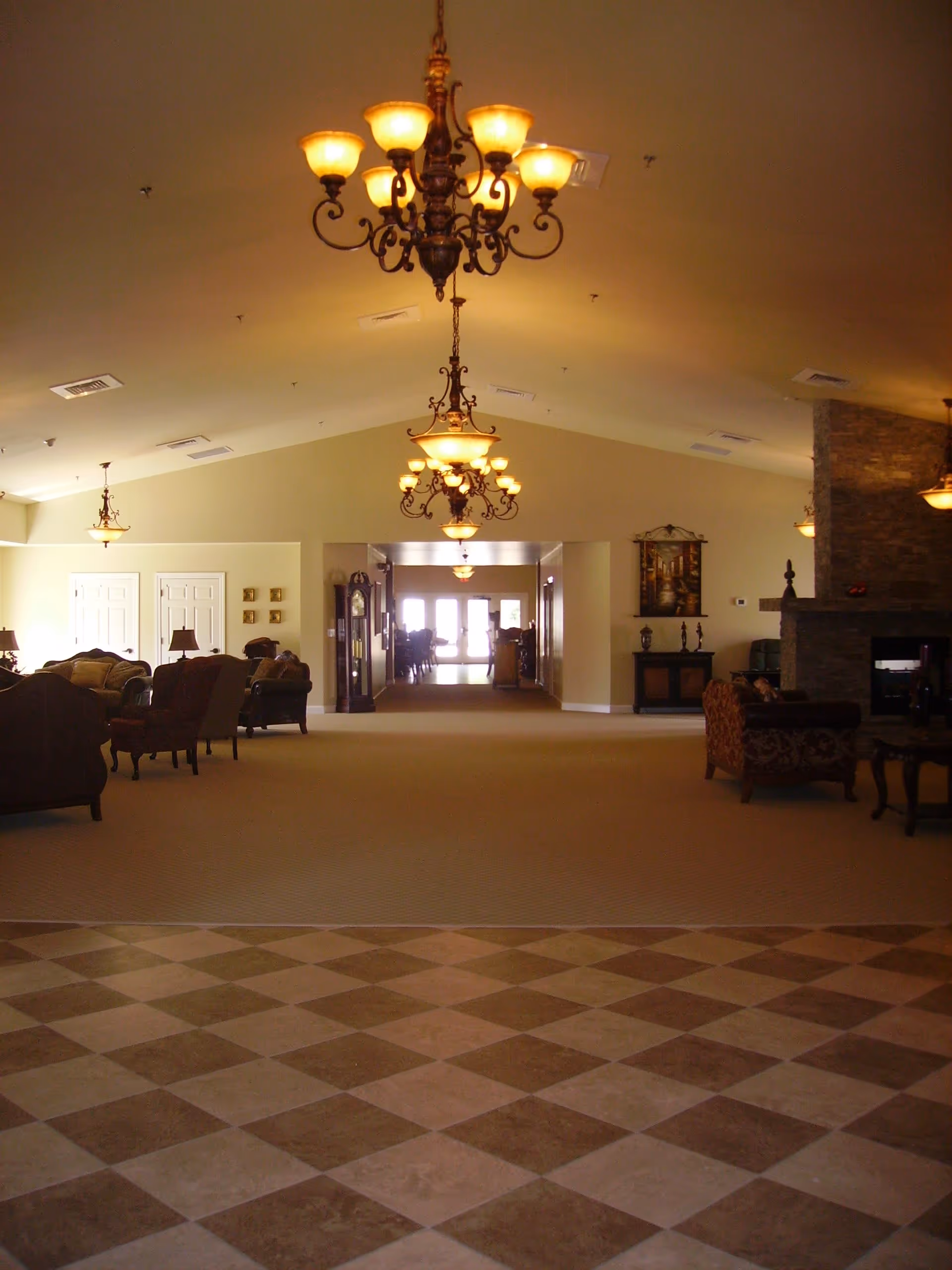 Spacious interior common area with patterned tile flooring in the foreground and carpeted seating area with sofas and armchairs in the background. Multiple ornate chandeliers hang from the high ceiling, and there is a stone fireplace on the right side. The room is warmly lit and has a welcoming atmosphere.
