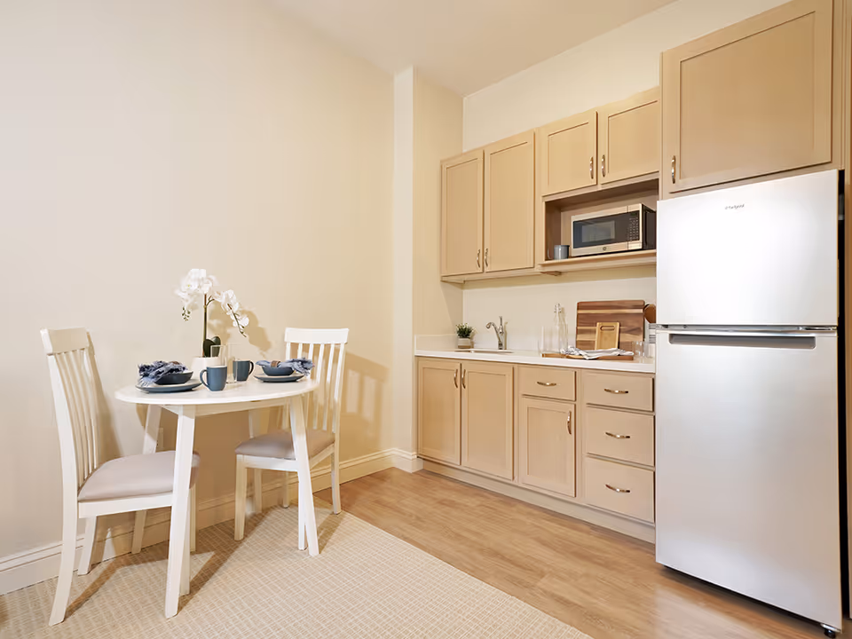 Small kitchenette with light wood cabinets, a stainless refrigerator, microwave, sink, and a round white dining table set for two.