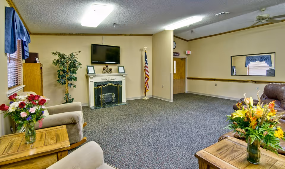 Common living room with upholstered chairs, side tables with flower arrangements, a fireplace with wall-mounted TV, and an American flag.