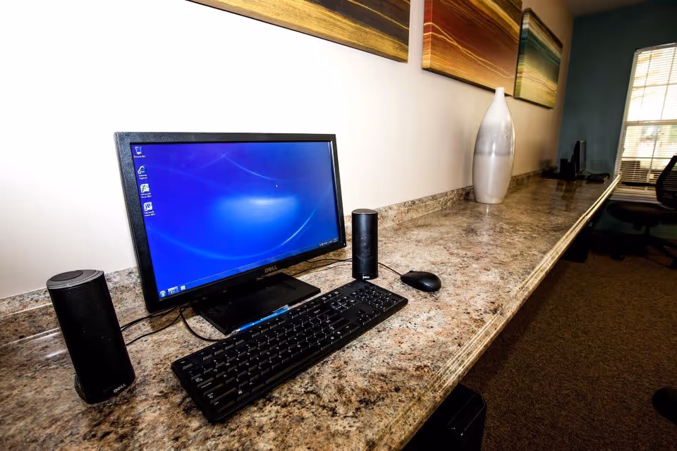 A computer workstation with a Dell monitor, keyboard, mouse, and two speakers on a long granite countertop. There is a large decorative vase and colorful abstract paintings on the wall above the countertop. A window with blinds and an office chair are visible in the background.