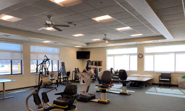 A spacious rehabilitation or physical therapy room with exercise equipment including stationary bikes and other fitness machines. The room has large windows letting in natural light, a ceiling fan, a wall-mounted TV, a therapy bed, chairs, and shelves with colorful therapy bands or equipment.