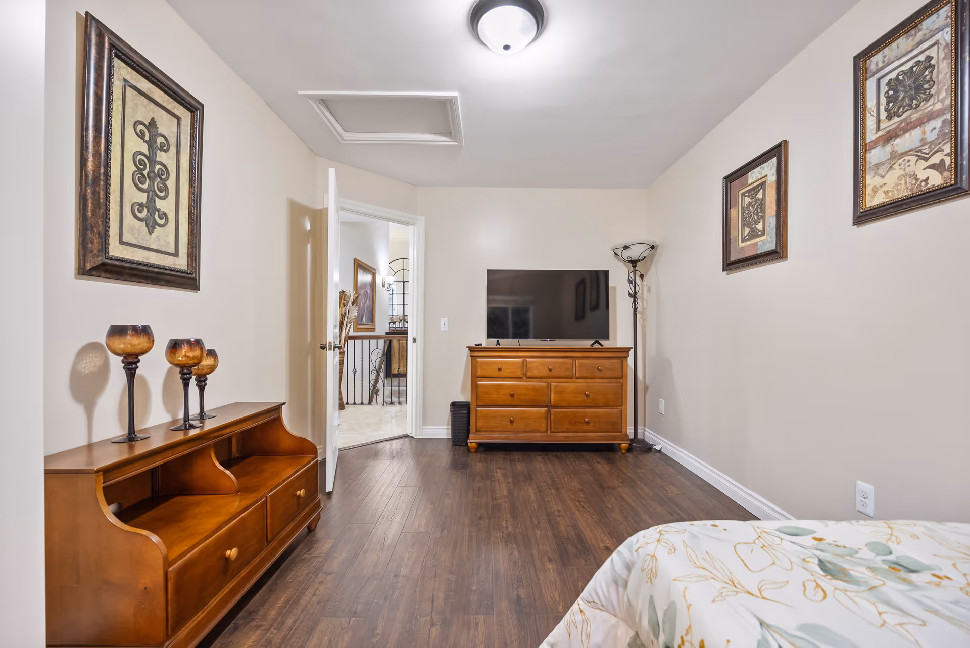A bedroom with wooden dressers, a TV, wall art, a floor lamp, and a partial bed visible in the foreground.