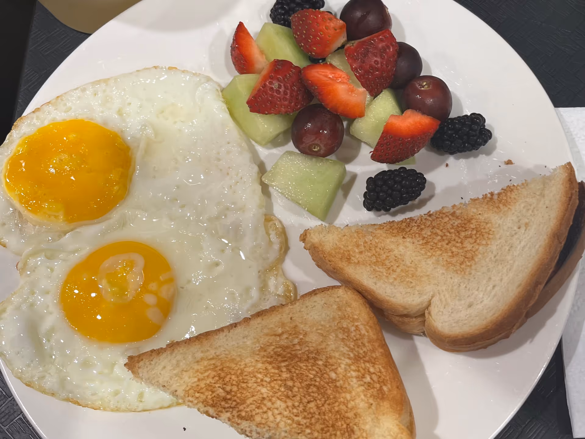 A white plate with two sunny-side-up eggs, two triangular pieces of toasted bread, and a small serving of mixed fresh fruit including strawberries, grapes, honeydew melon, and blackberries.