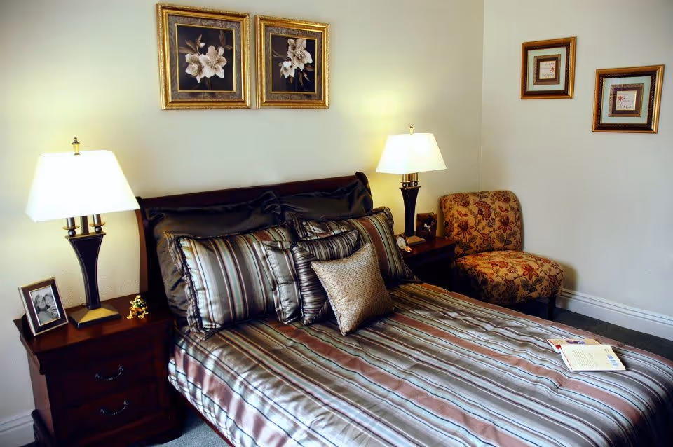 A neatly made bed with striped bedding and multiple pillows in a bedroom. There are two bedside tables with lamps, a framed photo, and a small decorative item. A floral-patterned chair is placed in the corner, and four framed pictures hang on the walls.