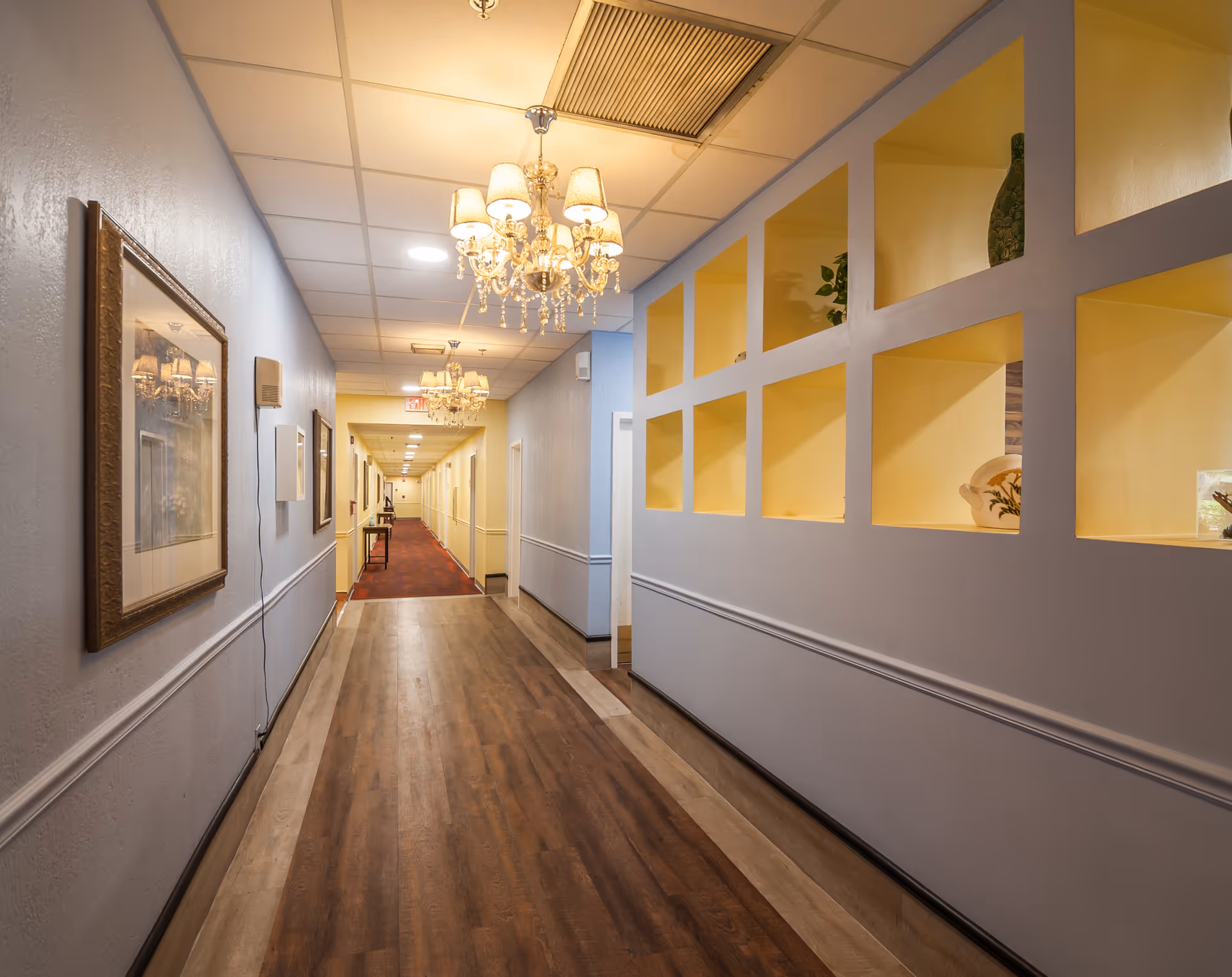 A well-lit hallway in a senior living facility with wooden flooring, light blue walls, and yellow recessed shelves displaying decorative items. The ceiling has chandeliers and recessed lighting, and framed artwork is hung on the left wall.