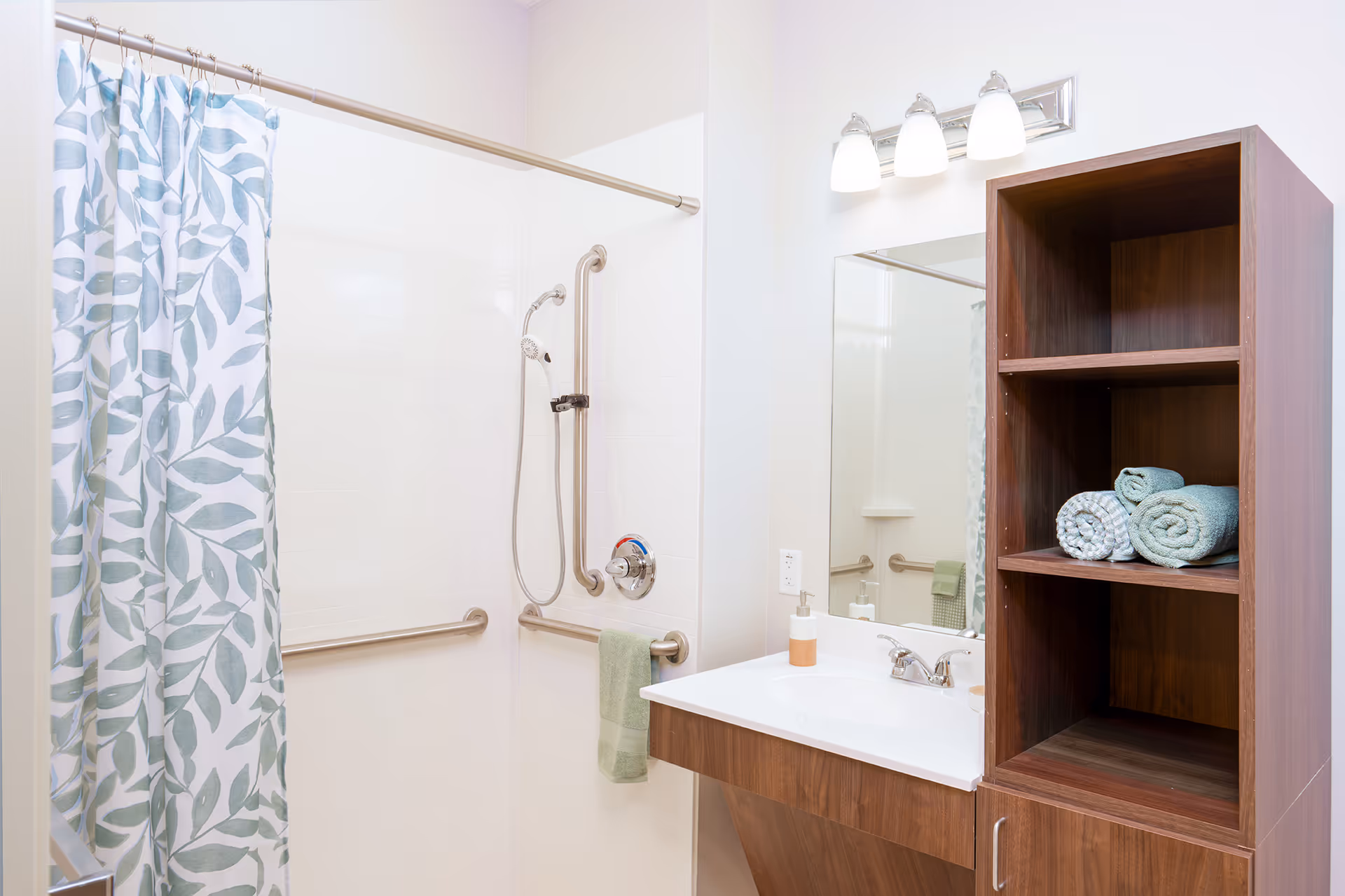 A clean and bright bathroom featuring a shower with a white and green leaf-patterned curtain, grab bars for safety, a handheld showerhead, a wooden vanity with a white sink, a soap dispenser, a large mirror, and a wooden shelving unit with rolled towels.