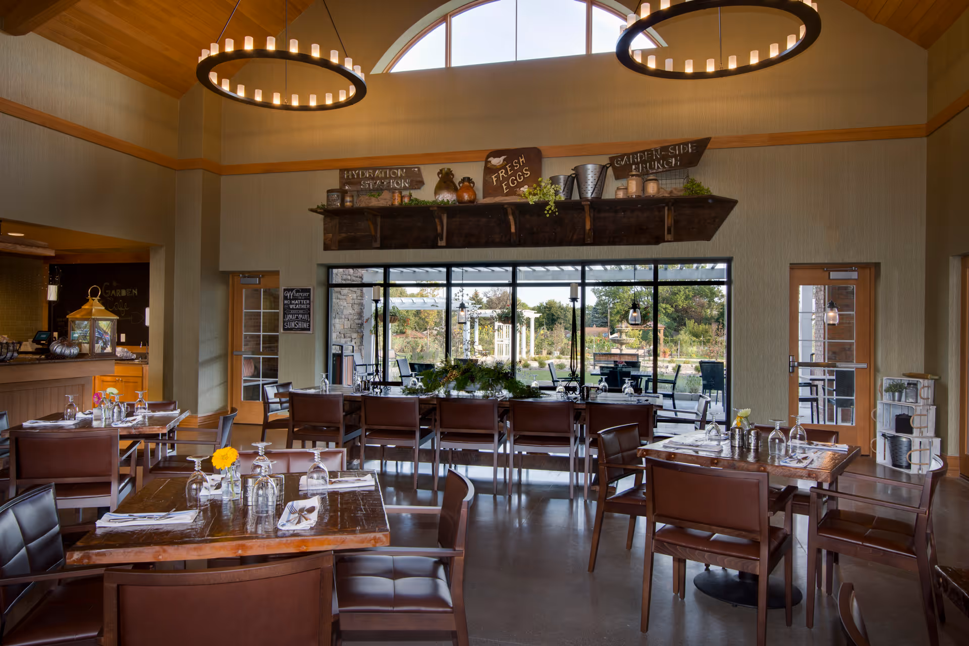 Interior view of a dining room with wooden tables and brown leather chairs. The tables are set with glasses, napkins, and silverware. Large windows and glass doors provide a view of an outdoor garden area. Two circular chandeliers hang from the ceiling, and decorative signs are displayed on a shelf above the windows.