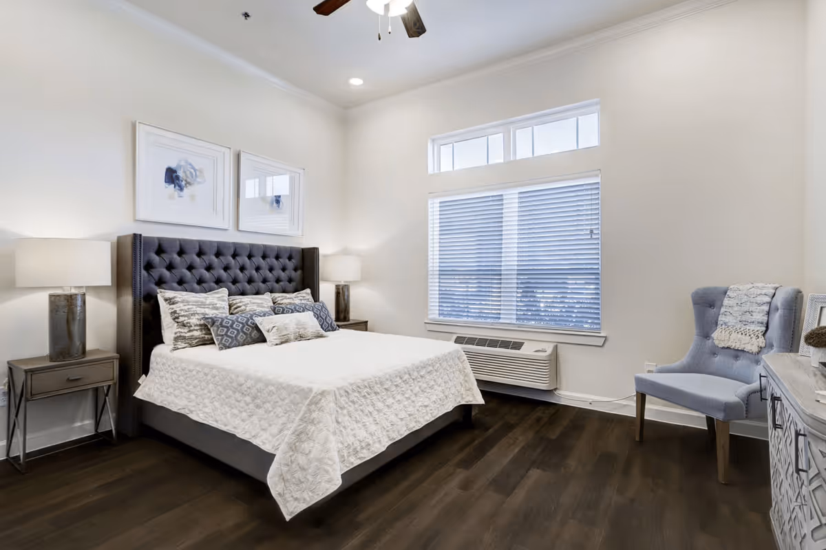 A neatly made bed with a tufted dark gray headboard and multiple decorative pillows in a bright bedroom. There are two framed abstract artworks above the bed, two bedside tables with lamps, a window with blinds, a blue upholstered chair with a throw blanket, and a dresser with decorative items. The room has dark wood flooring and white walls.