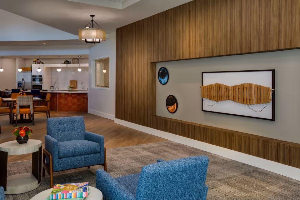 A modern living room area in a senior living facility featuring two blue upholstered armchairs, a round side table with a small flower arrangement, and a coffee table with books. The background shows a dining area with a table and chairs, pendant lights, and a kitchen with stainless steel appliances. The wall has a wooden panel with decorative art pieces mounted on it.