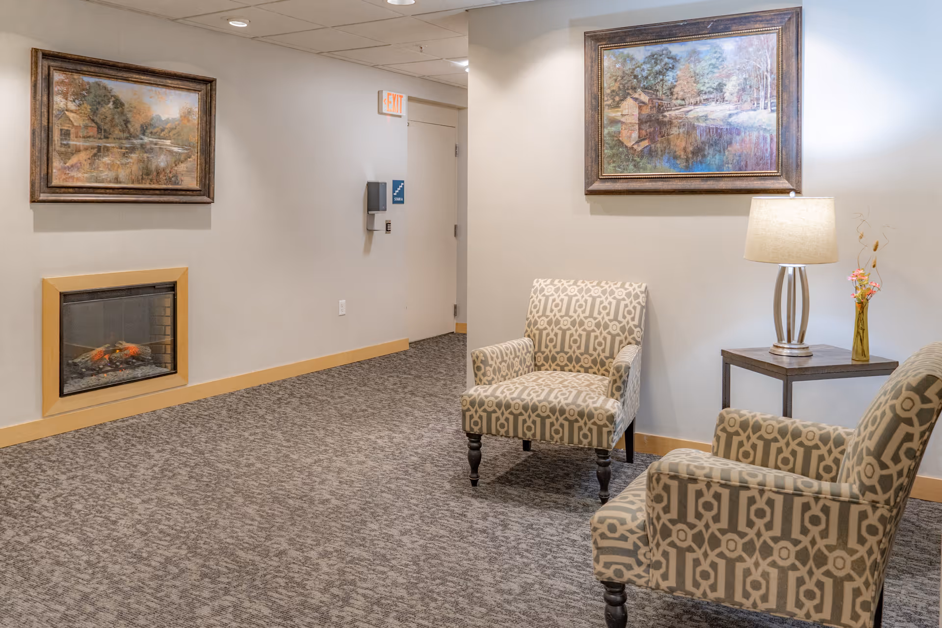 A cozy sitting area in a senior living facility with two patterned armchairs, a small wooden side table with a lamp and a vase with flowers, a wall-mounted electric fireplace, and two framed landscape paintings on the walls.