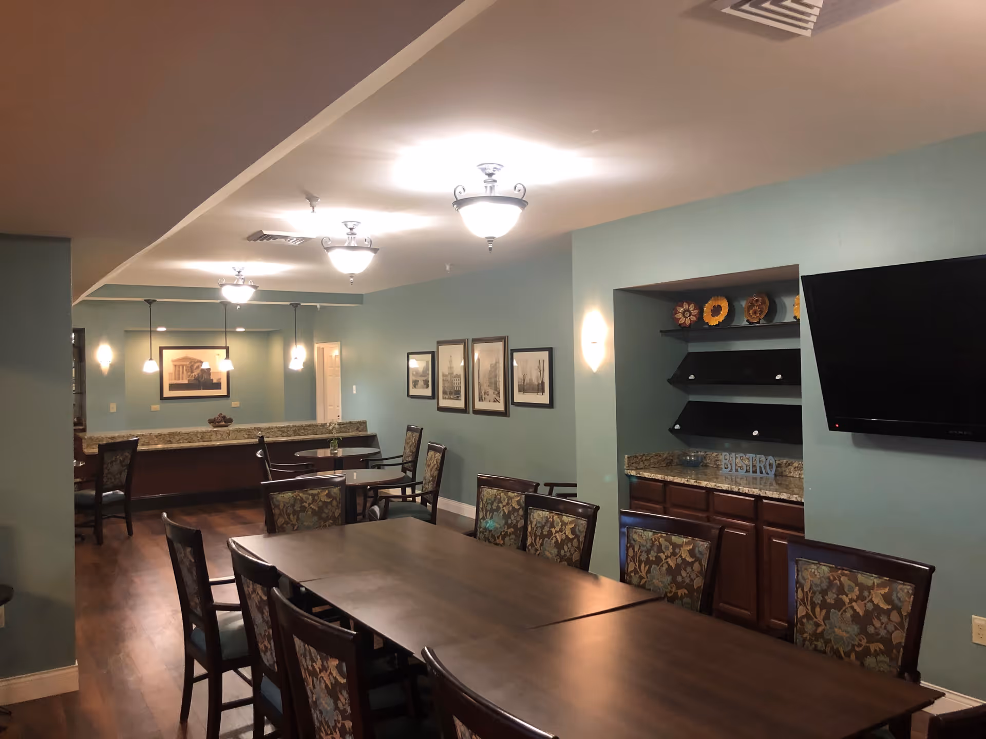 Interior dining room with a long wooden table surrounded by patterned chairs, a granite buffet counter, shelving with decor, and a wall-mounted TV.