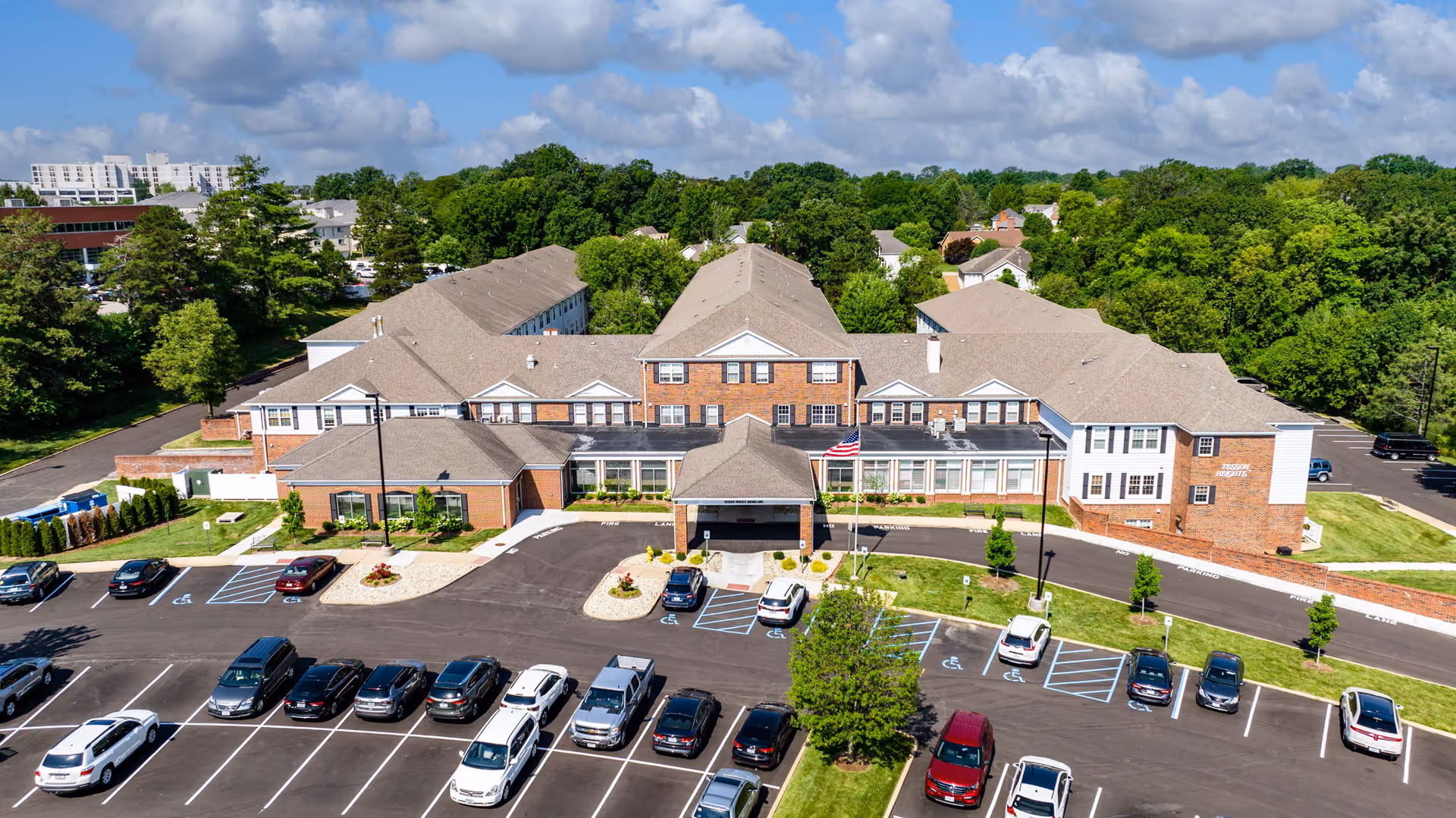 Aerial view of Cedarhurst Senior Living of Tesson Heights showing a large brick and beige building with multiple roof sections surrounded by a parking lot with several cars parked. The building is set against a backdrop of green trees and a partly cloudy sky.