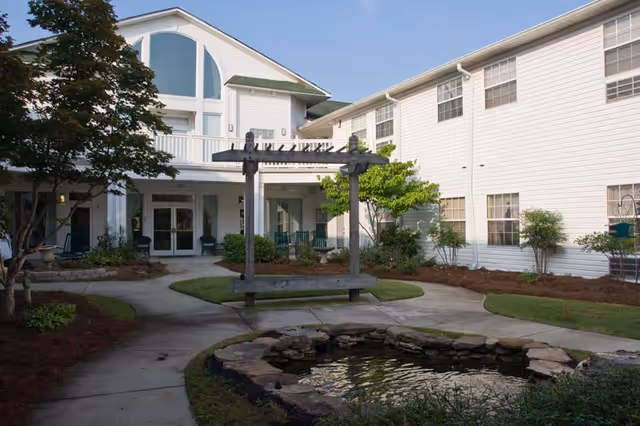 Outdoor courtyard area of a senior living facility with a white two-story building surrounding it. The courtyard features a small pond with rocks around it, a wooden pergola with a bench underneath, trees, shrubs, and a concrete walkway.