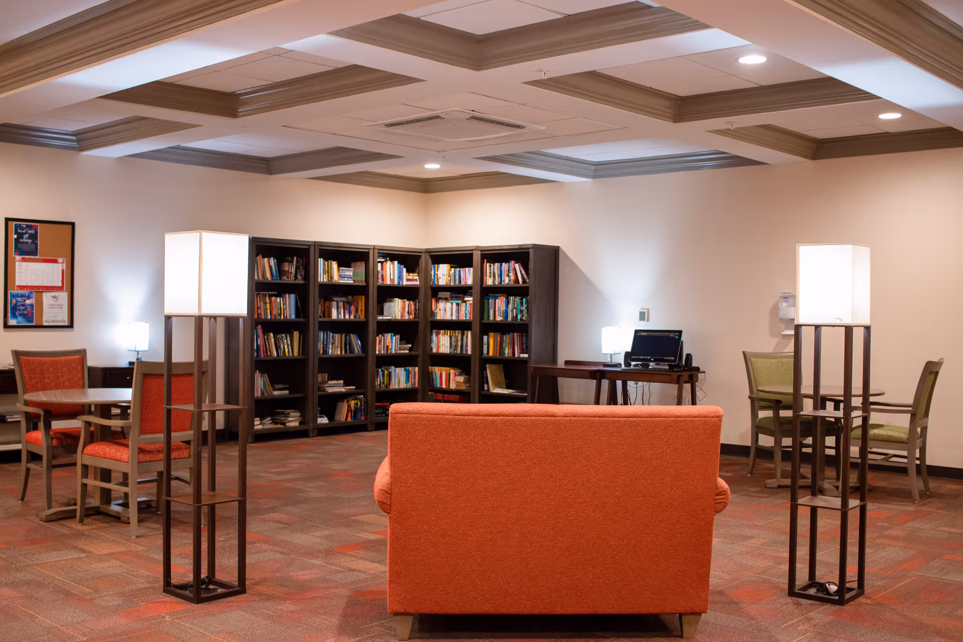 A cozy reading and sitting area in a senior living facility featuring a red couch facing away, two tall floor lamps with white shades, several bookshelves filled with books, and multiple tables with chairs in red and green upholstery. A computer desk with a monitor and lamp is visible against the wall.