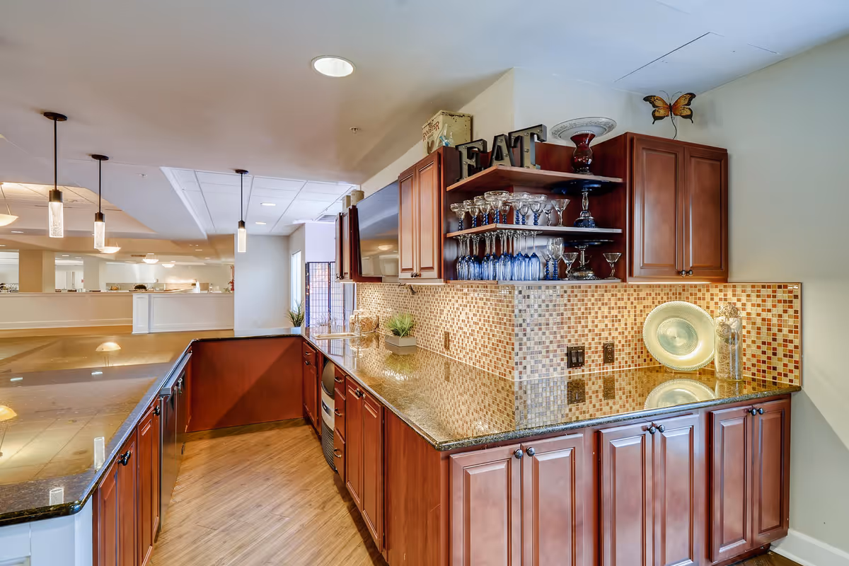 Open kitchenette/bar area with wooden cabinets, granite countertops, a mosaic tile backsplash and shelves of glassware overlooking a common lounge.