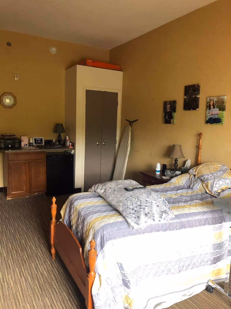 A bedroom in a senior living facility with a wooden bed featuring a patterned quilt and a folded blanket on top. There is a nightstand with a lamp, clock, and other small items next to the bed. On the wall above the nightstand are three framed photos. Across from the bed is a small cabinet with a mini fridge underneath, a lamp, and a digital clock showing the time 1:57. An ironing board is propped against the wall near a closed closet with double doors. The room has beige walls and carpeted flooring.
