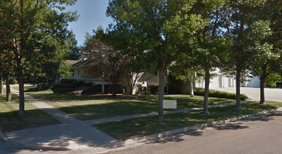 Residential-style senior facility exterior with a manicured lawn, trees, and a sidewalk.