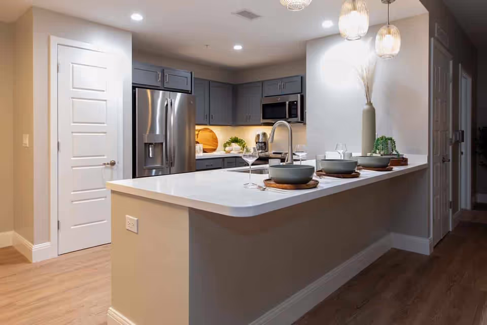 Modern kitchen with gray cabinets, stainless steel refrigerator and microwave, white countertops, and a breakfast bar set with bowls and glasses. Pendant lights hang above the bar, and there is a decorative vase with dried plants on the counter.