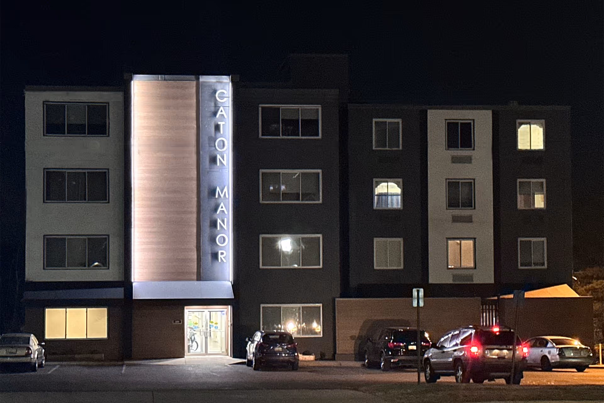 Nighttime view of a four-story apartment-style building with an illuminated vertical sign reading 'Caton Manor' and cars parked in front.