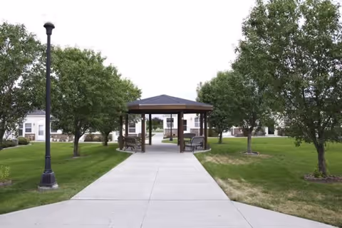 A paved walkway leads to a wooden gazebo with benches on either side, surrounded by green grass and trees in an outdoor area of a retirement community.
