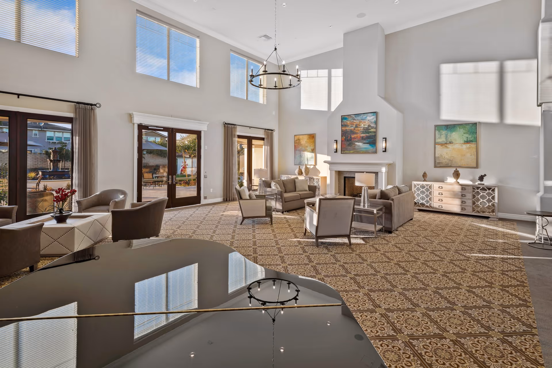 Spacious and well-lit living room area with high ceilings, large windows, and glass doors leading outside. The room features a patterned carpet, a grand piano in the foreground, multiple seating arrangements with sofas and chairs around a fireplace, decorative paintings on the walls, and a chandelier hanging from the ceiling.