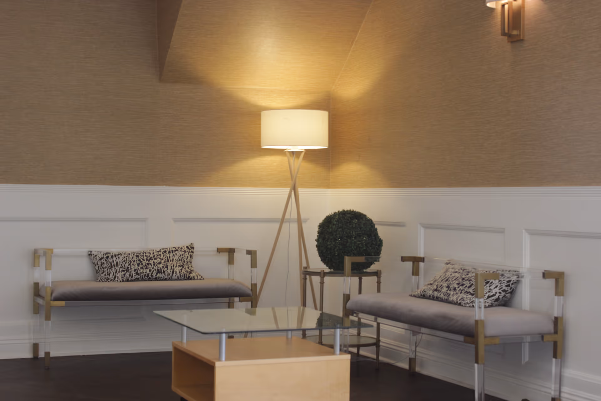 A cozy sitting area with two modern benches featuring gray cushions and patterned pillows, a glass coffee table with a wooden base, a small side table with a round green plant, and a tall floor lamp with a white shade illuminating the beige and white paneled walls.
